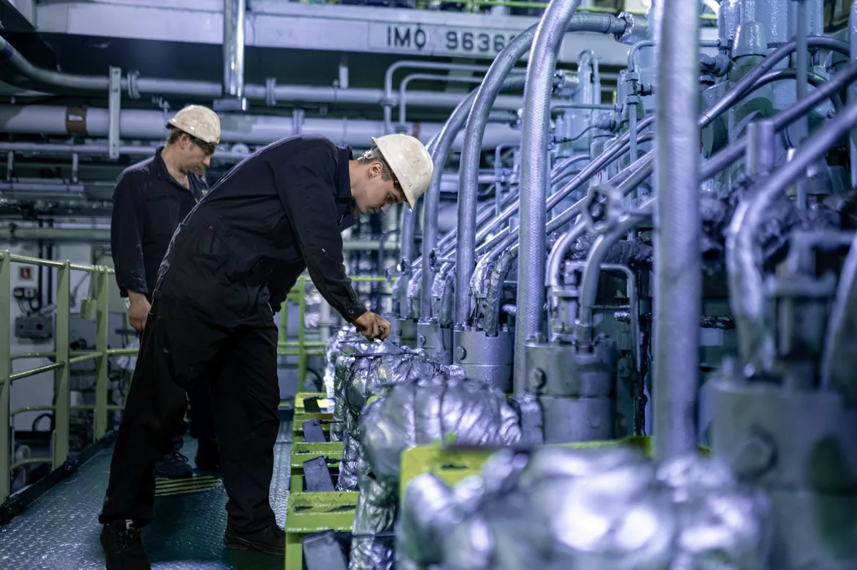 Two crew members in hard hats and overalls inspect a large marine engine with multiple cylinders and pipes in an engine room.