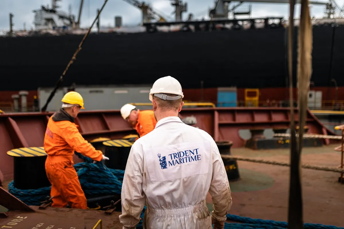 Three crew members, wearing jumpsuits and hard hats, are managing mooring lines on a ship's deck, with a large vessel docked behind them.