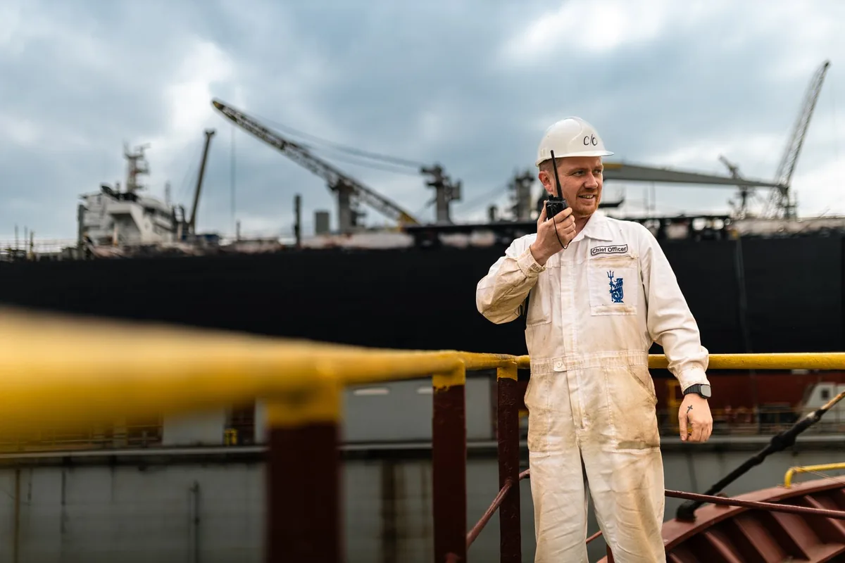 A Chief Officer in a white uniform and hard hat uses a walkie-talkie in a shipyard with a large vessel and cranes in the background.